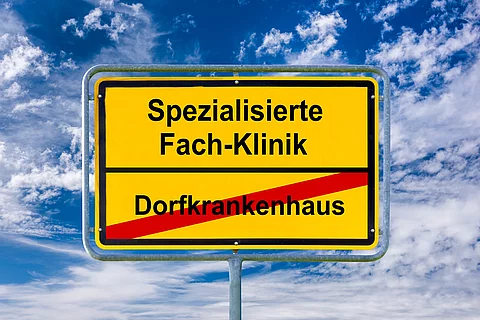 Ein Ortsschild ist beschriftet mit Dorfkrankenhaus und Spezialisierte Fach-Klinik - FOTOMONTAGE Ein Ortsschild ist beschriftet mit Dorfkrankenhaus und Spezialisierte Fach-Klinik - FOTOMONTAGE