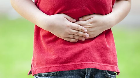 Ein Kind trägt ein rotes T-Shirt und blaue Jeans und hält sich mit beiden Händen den Bauch, was auf Bauchschmerzen hinweist.